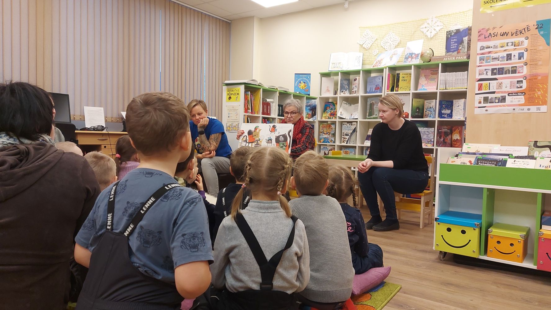 Bērni no Rīgas 154. pirmsskolas izglītības iestādes klausās, kā bibliotekāres lasa grāmatu "Runcis Puncis un bibliotēkā nenodotā grāmata".