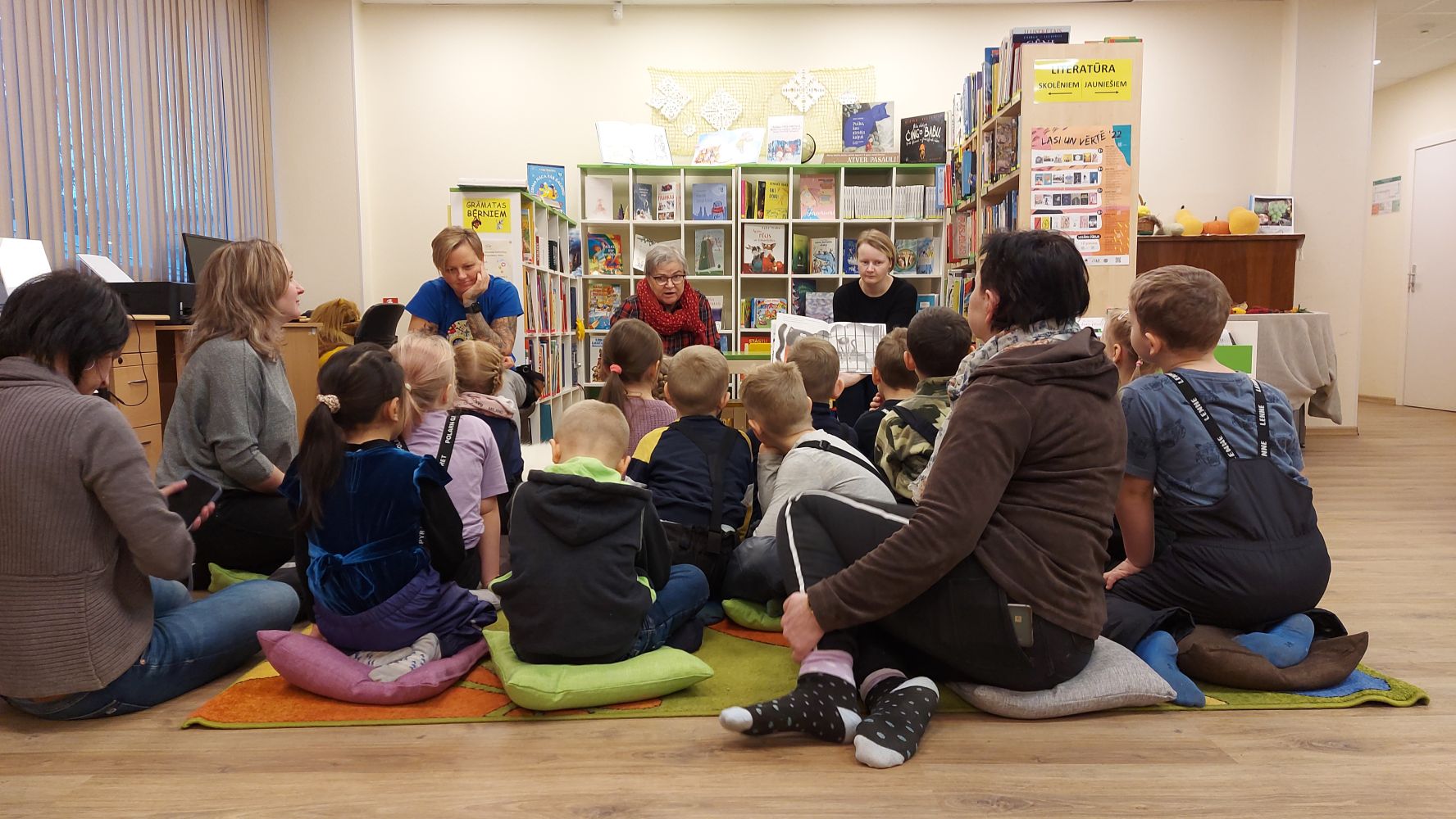 Bērni no Rīgas 154. pirmsskolas izglītības iestādes klausās, kā bibliotekāres lasa grāmatu "Runcis Puncis un bibliotēkā nenodotā grāmata".